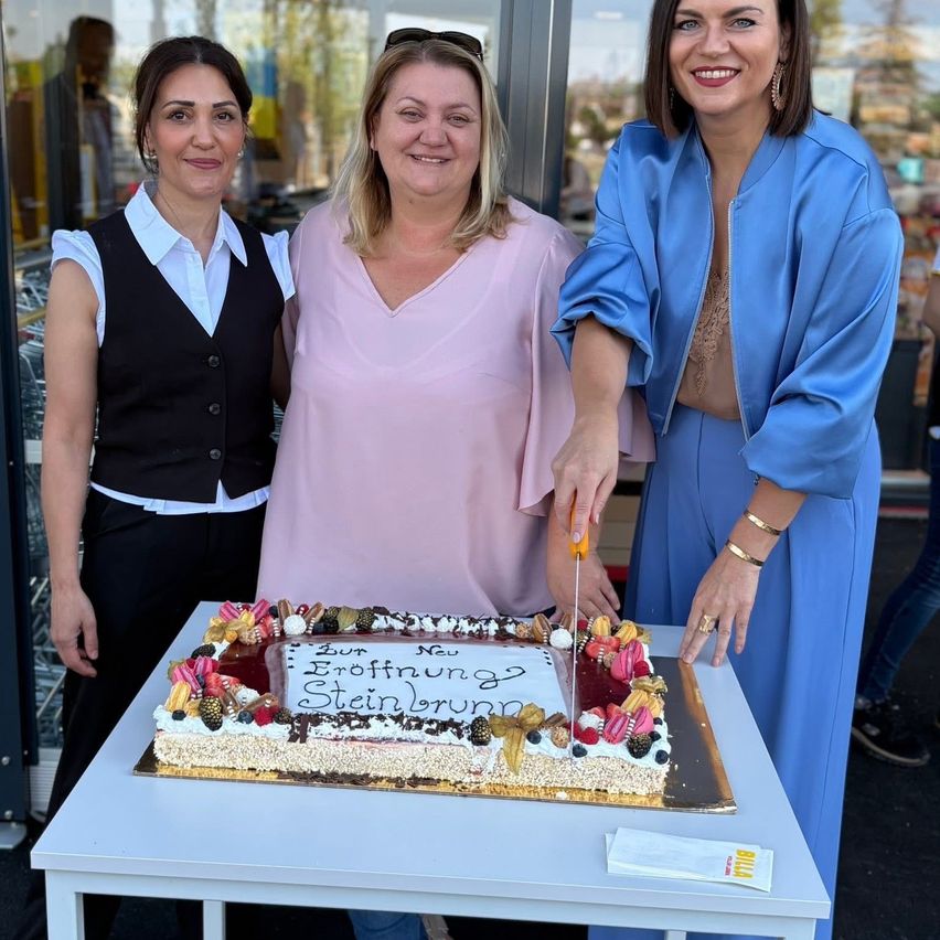 Drei Frauen stehen bei einem Tisch und schneiden einen Kuchen. Der Kuchen hat eine Nachricht in Deutsch geschrieben.