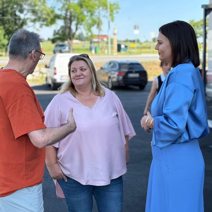 Drei Menschen stehen auf einem Parkplatz, eine Frau in Blau schaut zwei anderen gegenüber, einem älteren Mann in Orange und einer Frau in Rosa. Autos sind hinter ihnen geparkt.