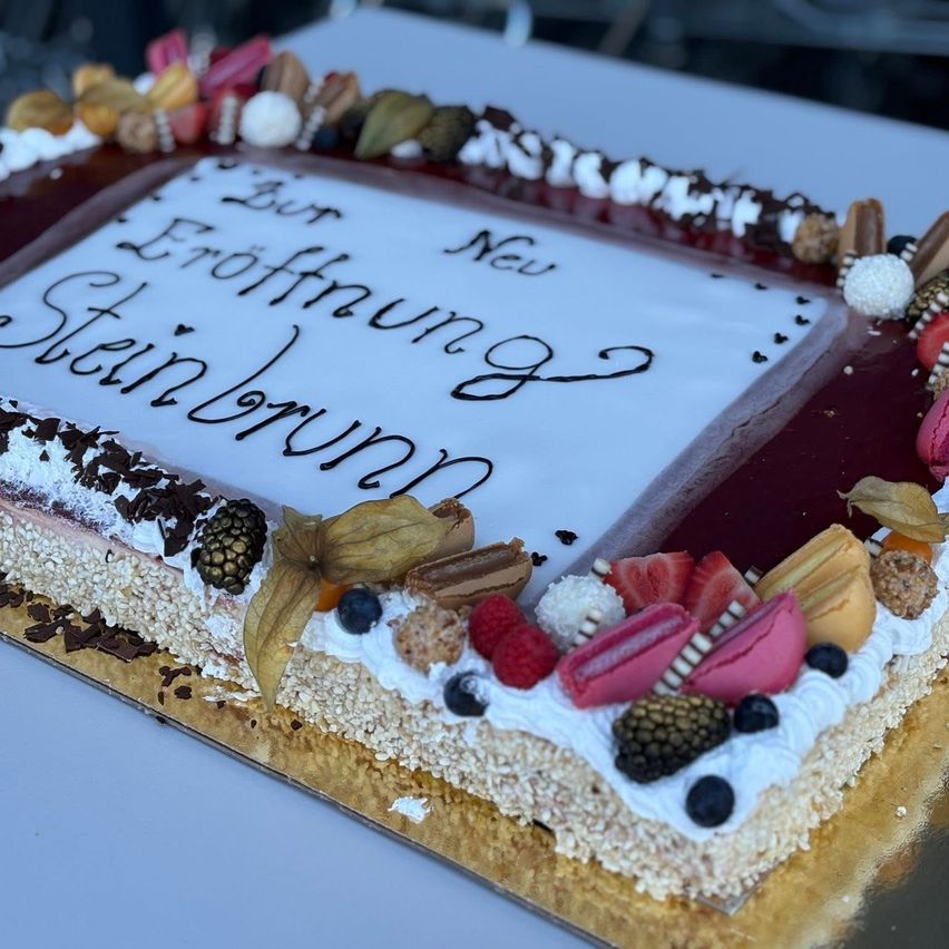Ein dekorativer Kuchen mit weißer Glasur, verziert mit verschiedenen Früchten und Süßigkeiten. Auf dem Kuchen stehen die Worte 'Gut Eroffnung Steinbrunnen'.