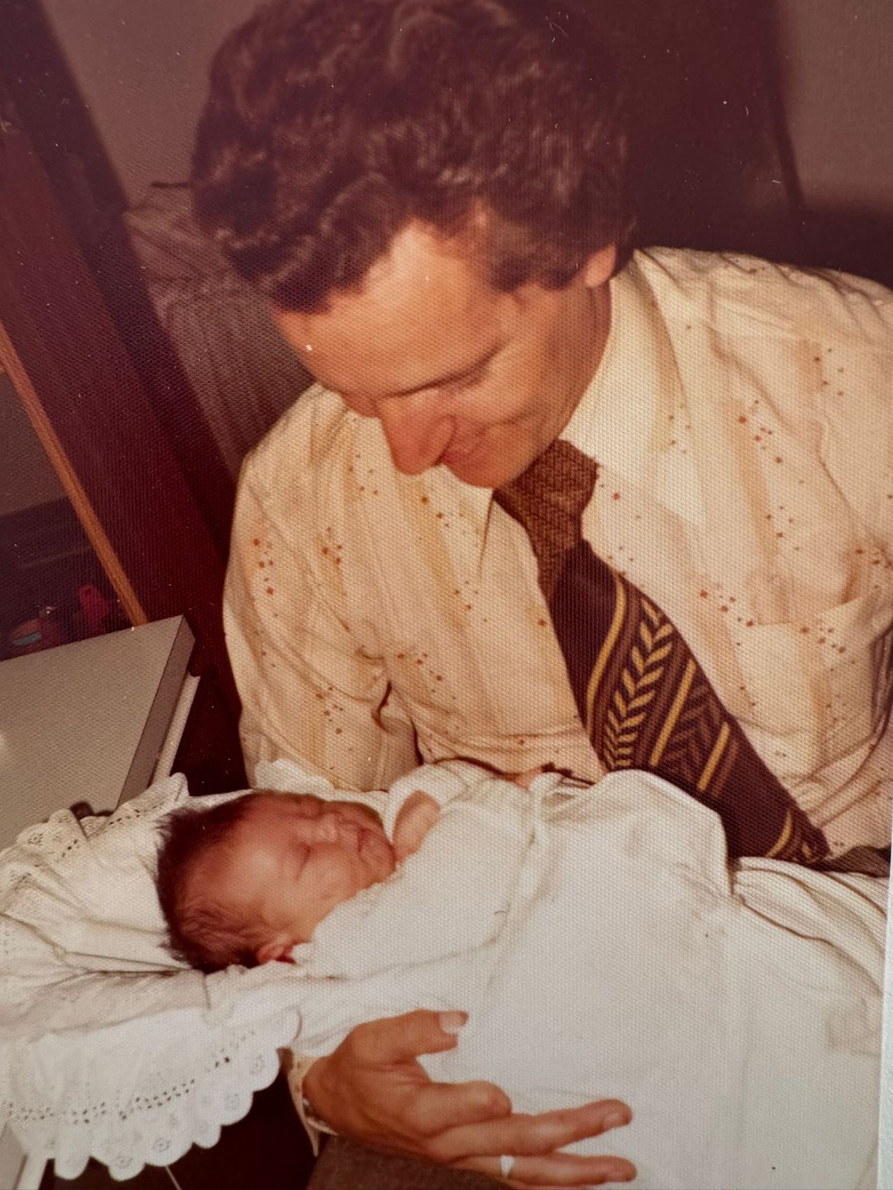 Ein Mann in einer Krawatte hält ein schlafendes Baby. Das Hemd des Mannes hat Flecken. Das Baby ist in eine Decke gewickelt.