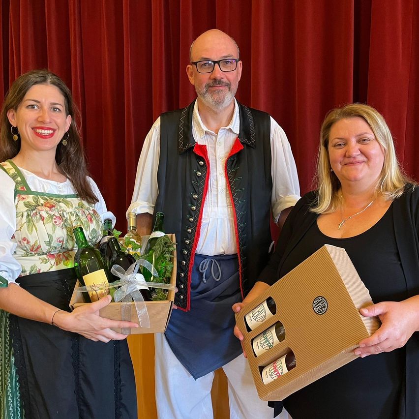 Drei Personen stehen vor einem Vorhang. Eine Frau hält eine Weinschachtel, ein Mann trägt eine traditionelle Kleidung und eine andere Frau hält eine Geschenkbox. Sie lächeln für ein Foto.