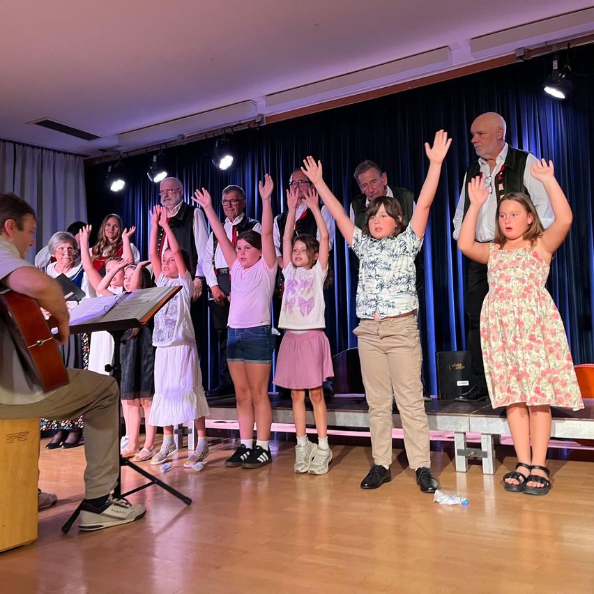 Eine Gruppe von Menschen, einschließlich Kinder, steht auf der Bühne und hebt die Hände, wahrscheinlich bei einer Aufführung. Ein Mann mit einer Gitarre sitzt auf einem Hocker links.