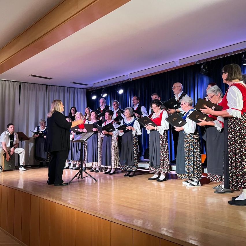 Ein Chor von Frauen in traditioneller Kleidung auf der Bühne mit einer Regisseurin vor ihnen, sie halten Notenbücher in den Händen, unter Bühnenbeleuchtung.
