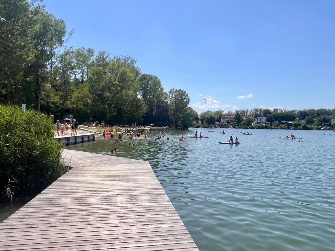 Ein ruhiger See mit vielen Menschen, die schwimmen und auf dem Wasser treiben. Ein hölzerner Steg erstreckt sich ins Wasser, und mehrere Bäume und Häuser umgeben das Gebiet.