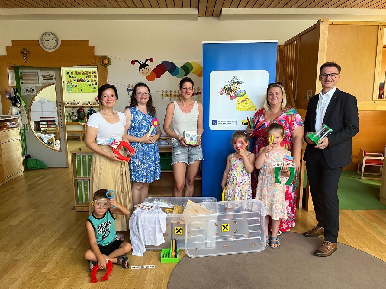 Eine Gruppe von Frauen und Kindern mit einem Mann posiert für ein Foto in einem Klassenzimmer mit Spielzeug und einem Banner im Hintergrund.