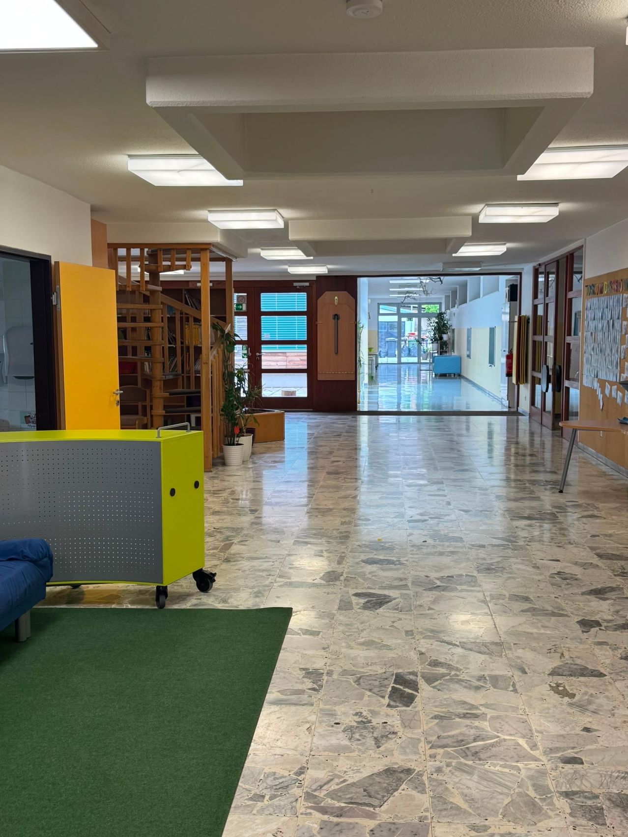 An empty hallway with marble floors, white walls, and yellow doors. Potted plants, a yellow cabinet, and a blue couch are on the left. The ceiling has several fluorescent lights.