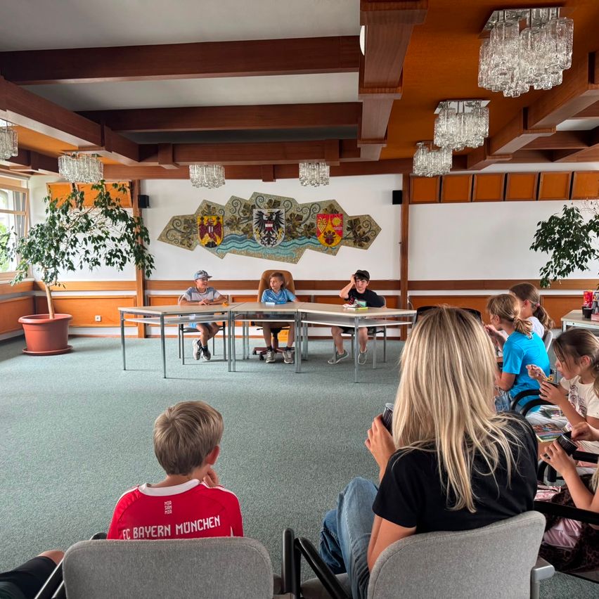 A group of people are sitting in chairs around a table in a room. A few children are sitting on the chairs. A woman is holding a camera.