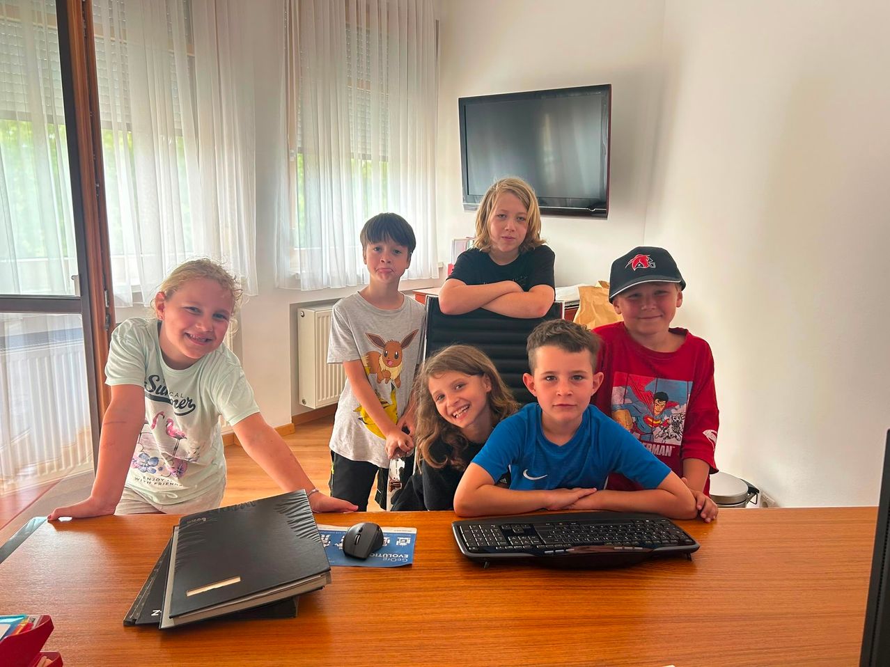 A group of children are sitting around a desk in a room. They are smiling and posing for a photo. There is a monitor, a radiator, and a bag behind them.