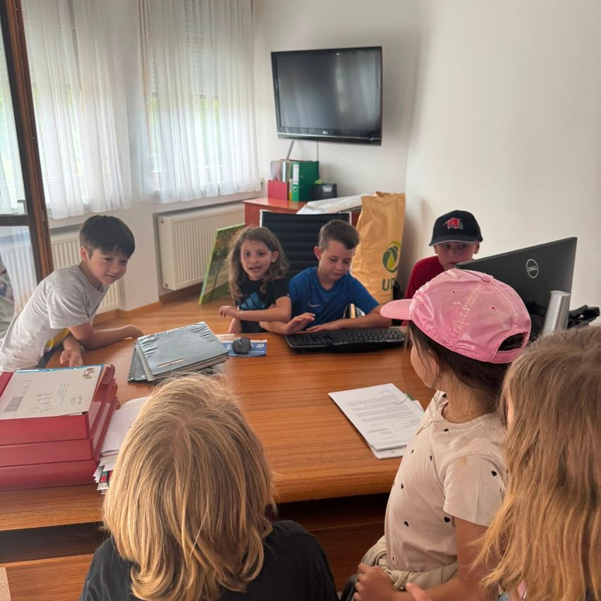 A group of children are sitting around a desk in a room. They are engaged in various activities, and a girl in a pink hat is looking at the camera.