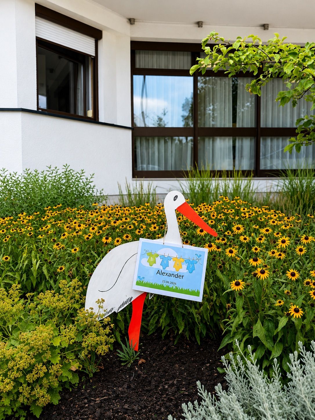 A stork with a sign that says Alexander is standing in a garden full of sunflowers in front of a white building.