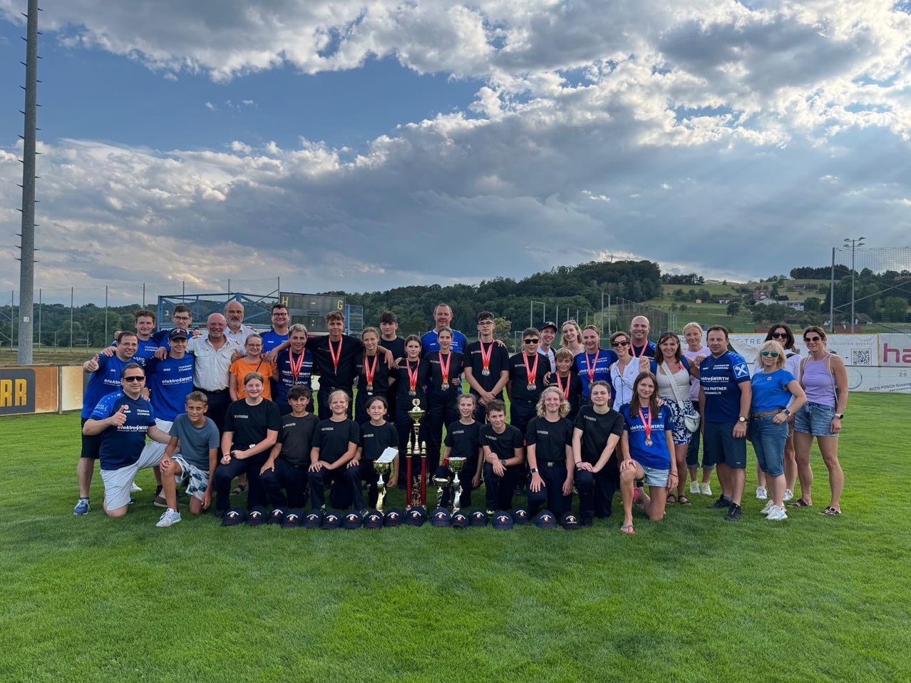 A group of people in matching blue shirts and hats are posing for a photo on a grassy field with trophies and medals.