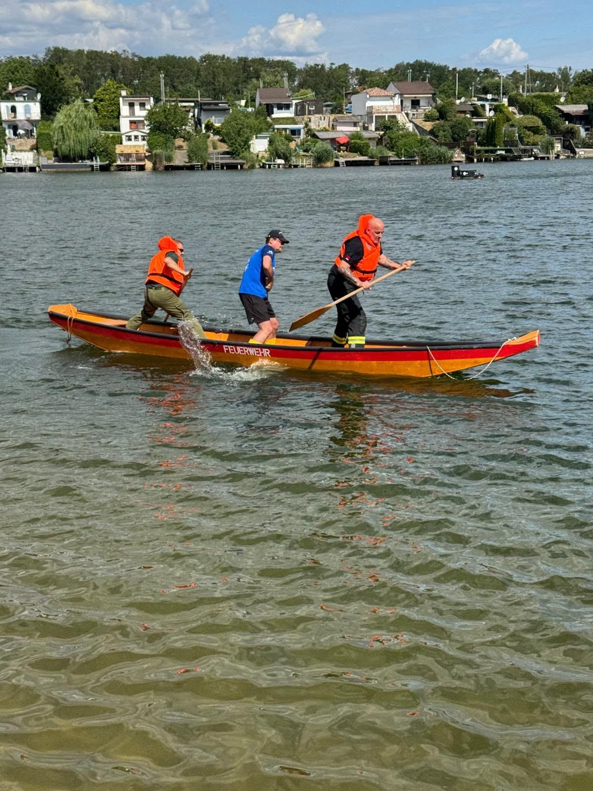 Three men in life jackets paddle a canoe on a calm lake, with houses and trees in the background.