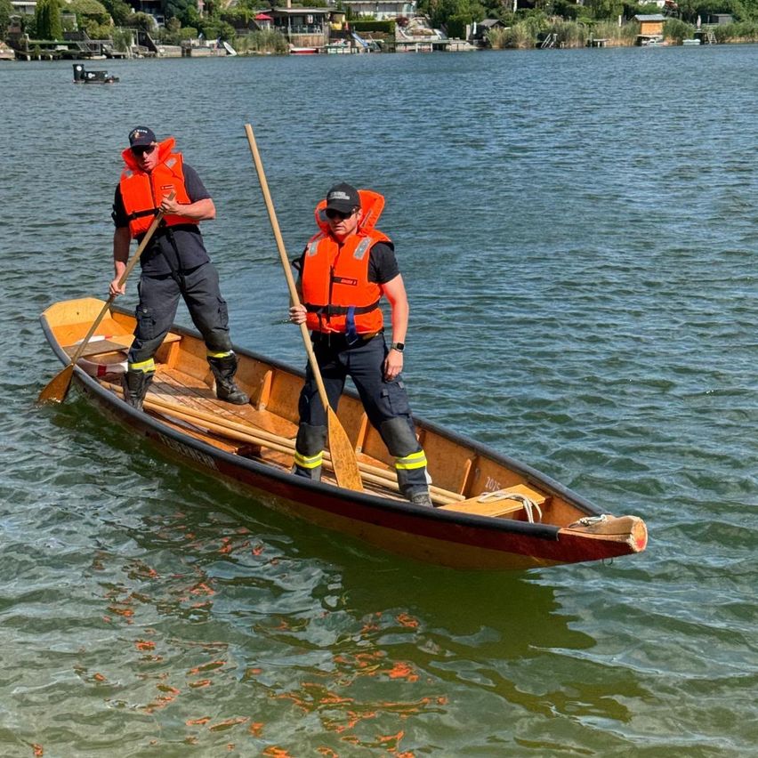 Two men in orange life jackets are standing in a small boat, holding paddles, and paddling on a calm lake.