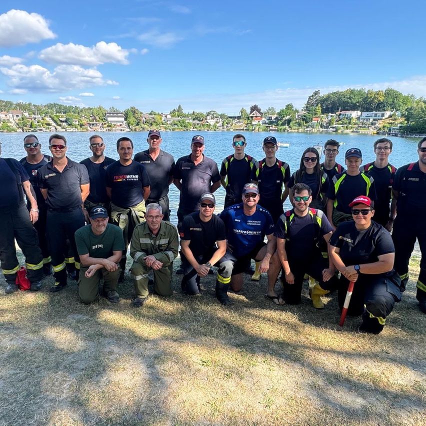 A group of firefighters in uniform pose for a photo by a lake with a backdrop of trees and houses.