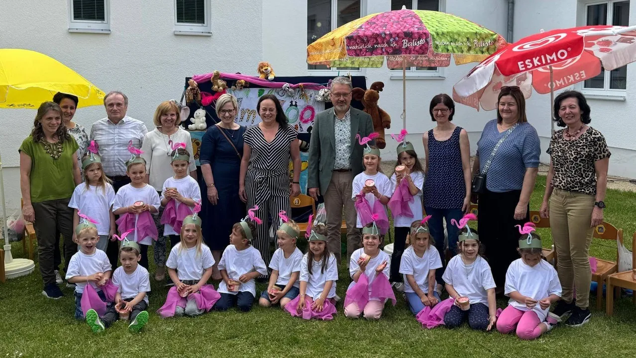 Eine Gruppe von Erwachsenen und Kindern versammelt sich auf einer Rasenfläche. Sie tragen rosa Hüte und halten Tassen. Ein Mann in einem grünen Jackett steht unter ihnen. Hinter ihnen befindet sich eine dekorierte Kulisse mit Plüschtieren und einem Regenschirm.