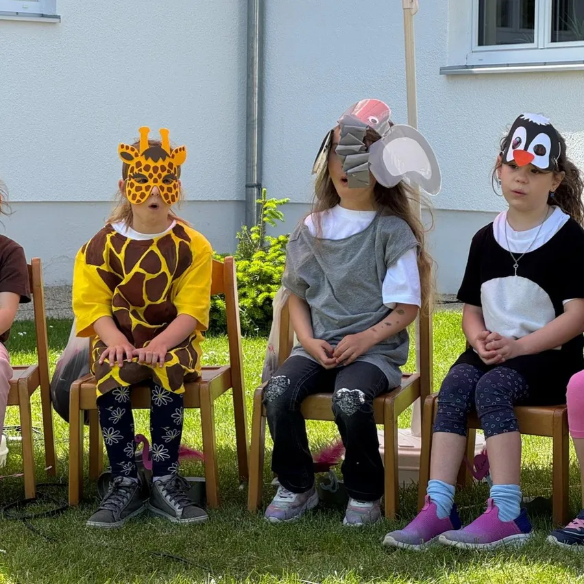 Drei Kinder in Tierkostümen sitzen auf Stühlen im Freien, eines als Giraffe, eines als Elefant und das dritte als Pinguin verkleidet. Dahinter steht ein weißes Gebäude mit einem Fenster.