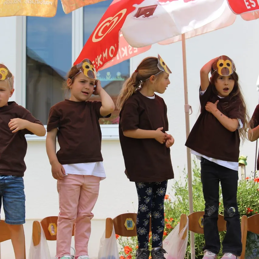Fünf Kinder in braunen T-Shirts und Tierkopfbedeckungen stehen unter einem Schirm, wobei zwei junge Mädchen ihre Ohren halten und ein Junge Turnschuhe trägt.