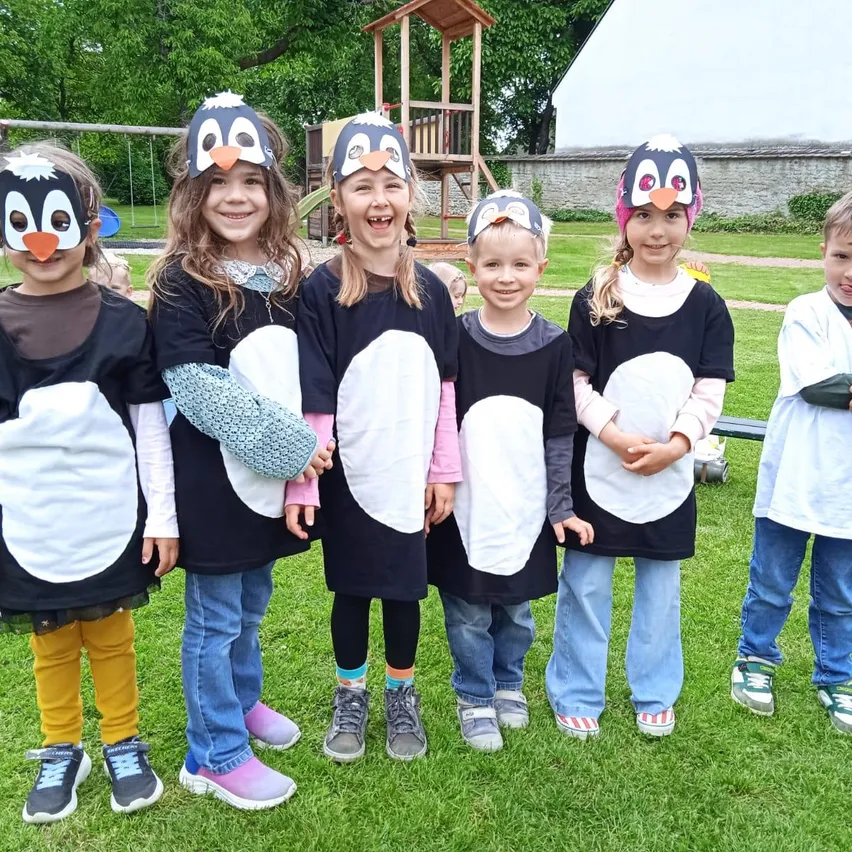 Eine Gruppe von Kindern in Pinguinkostümen, die auf einem Rasenfeld posieren, mit einem Spielplatz im Hintergrund.