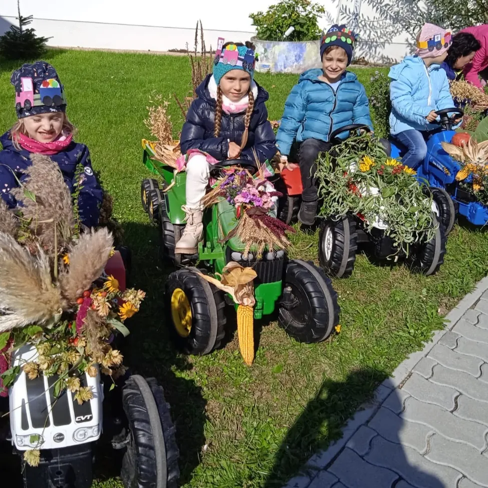 Mehrere Kinder sitzen auf Spielzeugtraktoren mit Blumendekorationen. Sie tragen Winterkleidung und Mützen. Im Hintergrund befindet sich ein Gebäude und ein Garten.