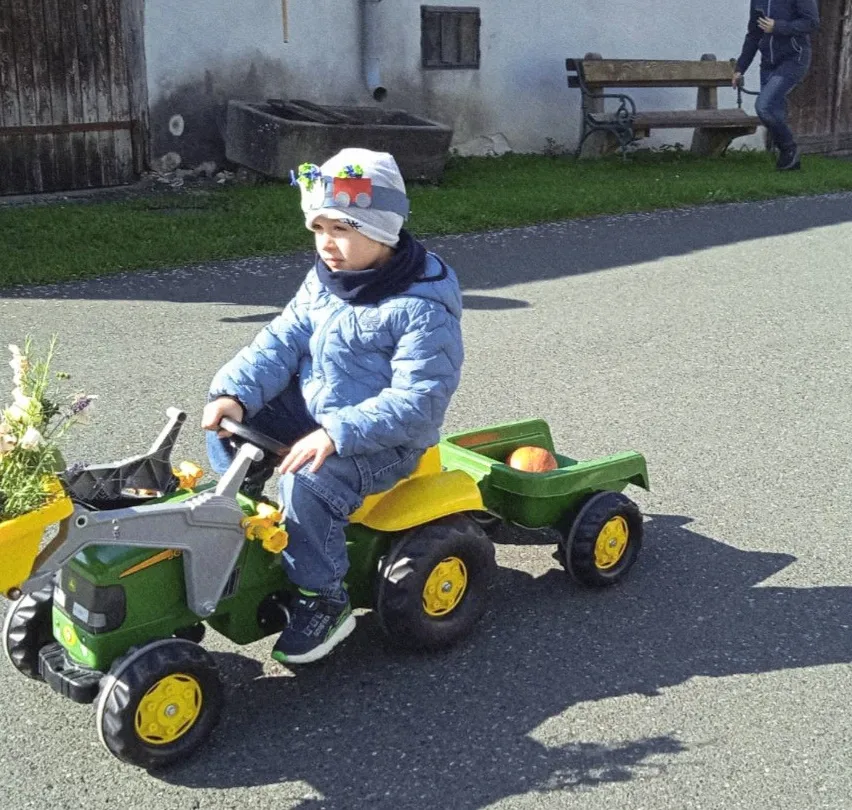 Ein junges Kind in einem Wintermantel fährt einen Spielzeugtraktor mit Blumen in einem gelben Eimer. Eine Person geht dahinter auf einem Weg.
