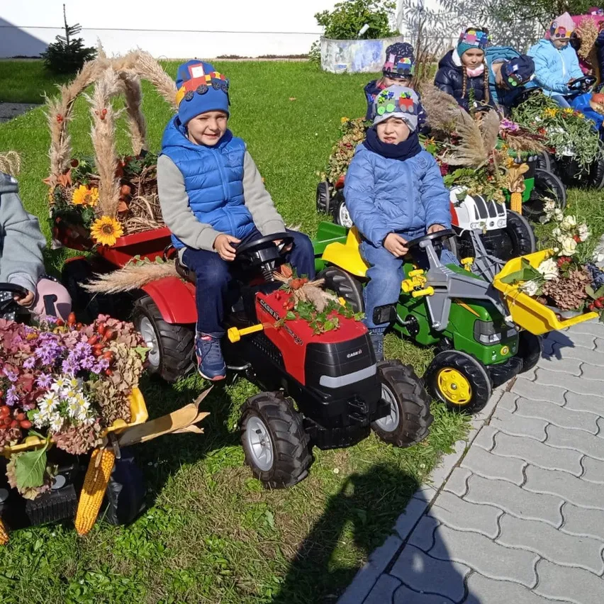 Kinder in Winterkleidung sitzen auf Spielzeugtraktoren auf einem Rasen. Die Traktoren sind mit Blumen und Pflanzen geschmückt. Der Boden ist mit Gras bedeckt.