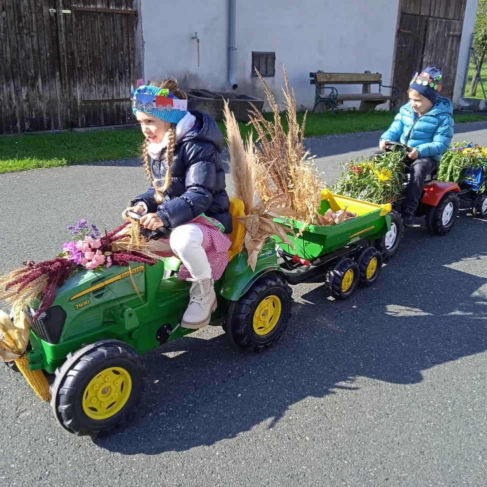Zwei Kinder in Winterkleidung fahren Spielzeugtraktoren mit Blumenarrangements in einer sonnigen Außenumgebung. Dahinter fährt ein weiteres Kind einen roten Traktor. Im Hintergrund steht ein Holzgebäude mit einer Bank.