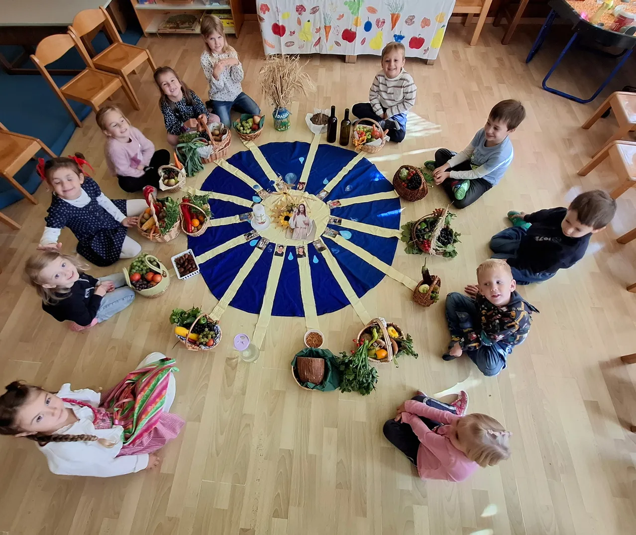 Kinder sitzen in einem Kreis mit Körben voller Obst und Gemüse. Ein blauer kreisförmiger Stoff ist in der Mitte mit einem Bild einer Frau. Einige Flaschen sind auf dem Tuch platziert.