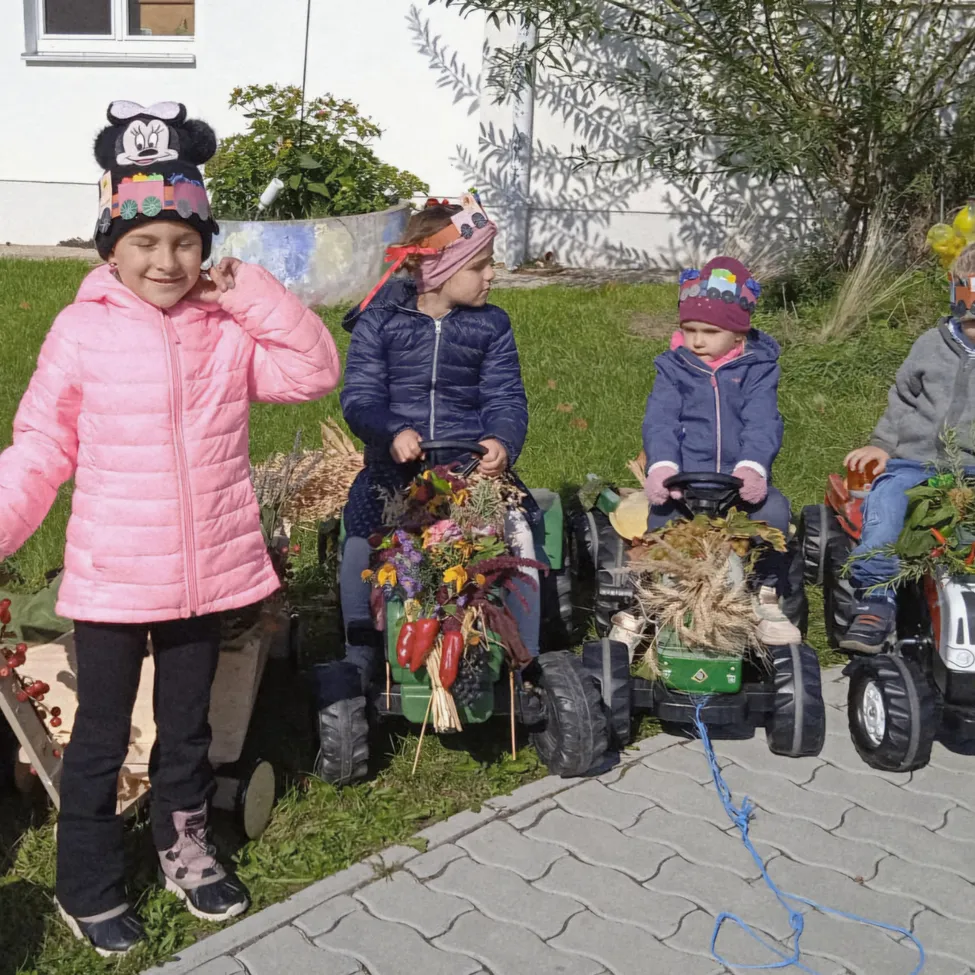 Vier Kinder in Winterkleidung sitzen auf Spielzeugtraktoren und ziehen Girlanden aus Herbstblättern. Ein Mädchen in Pink steht neben den Traktoren und lächelt. Sie sind in einem ruhigen Garten mit einem weißen Haus und Pflanzen im Hintergrund.