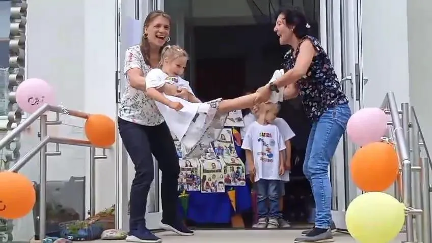 Zwei Frauen spielen mit einem kleinen Mädchen, halten es hoch und lachen. Das Mädchen trägt ein weißes Shirt. Hinter ihnen stehen zwei Kinder, und es gibt Luftballons und einen Tisch mit Bildern.