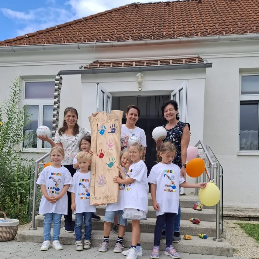 Mehrere Kinder und Erwachsene stehen auf den Stufen eines Hauses, lächeln und halten ein Holzbrett mit Handabdrücken und den Worten 'Vorschulkinder 2022'. Sie tragen weiße T-Shirts mit bunten Designs und halten Luftballons. Das Haus hat ein braunes Dach und weiße Wände.