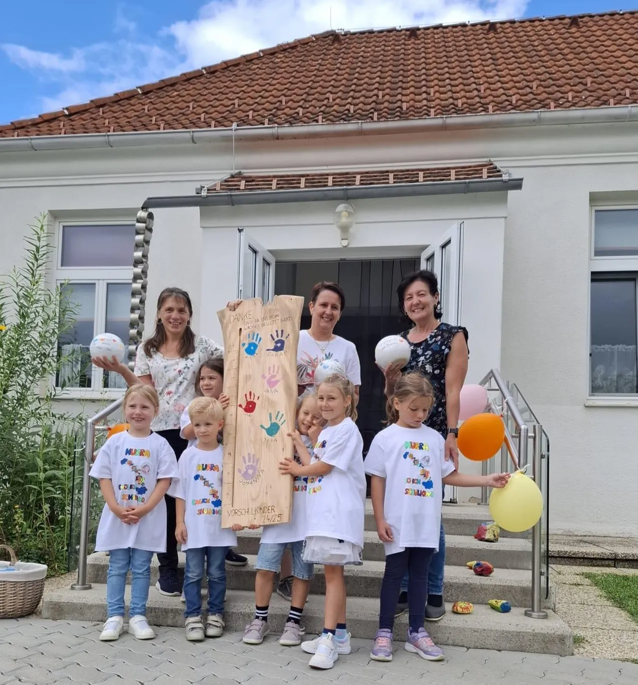 Eine Gruppe von Kindern und Erwachsenen steht vor einem Haus und hält ein Holzbrett mit Handabdrücken. Sie lächeln und scheinen für ein Foto zu posieren.