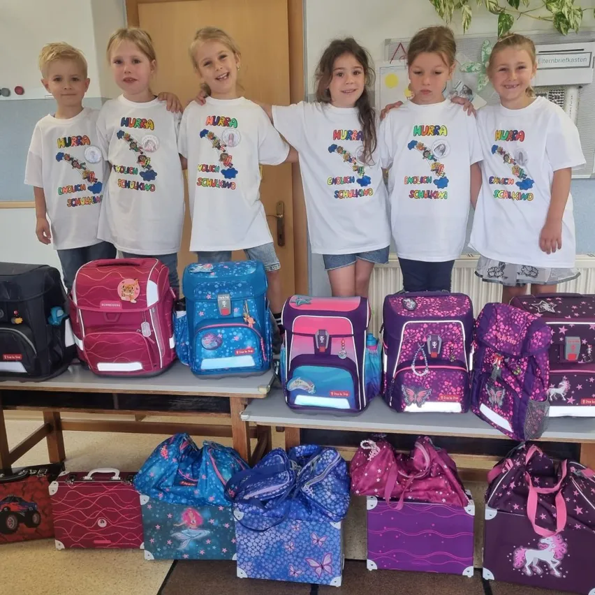 Eine Gruppe von Kindern, wahrscheinlich in einem Klassenzimmer, trägt weiße T-Shirts mit 'Murra' darauf. Sie stehen hinter einem Tisch, auf dem verschiedene farbenfrohe Rucksäcke und Taschen stehen. Die Kinder scheinen für ein Foto zu posieren.