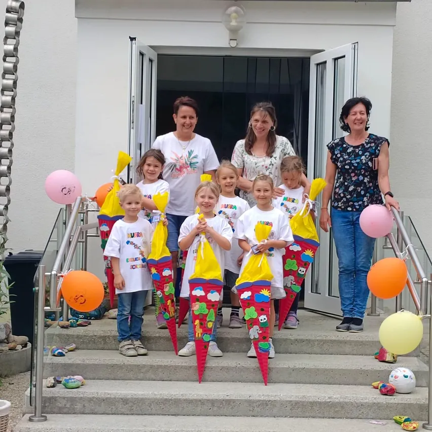 Eine Gruppe lächelnder Kinder und Erwachsener steht auf den Stufen eines Gebäudes und hält bunte Taschen. Sie sind von Luftballons umgeben und eine Kettengitterzaun befindet sich links.