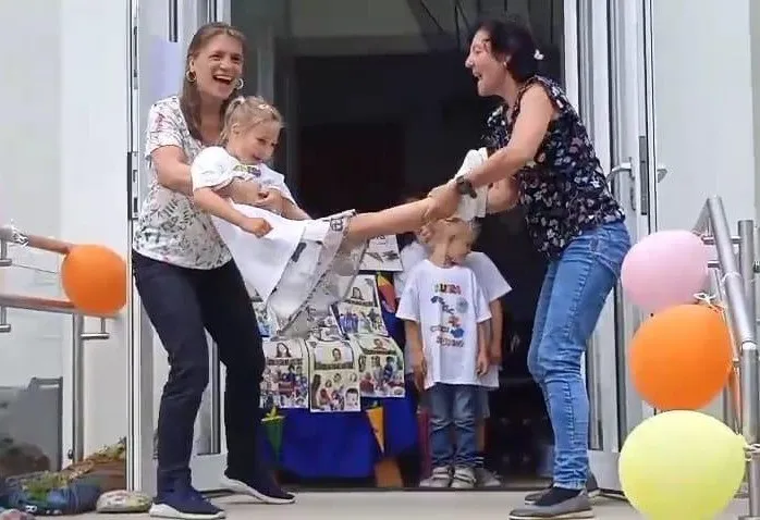 Zwei Frauen halten ein junges Mädchen, und sie tritt mit dem Bein. Das Mädchen trägt ein weißes Kleid. Hinter ihnen stehen zwei Kinder und beobachten. Bunte Luftballons sind an den Geländern befestigt.