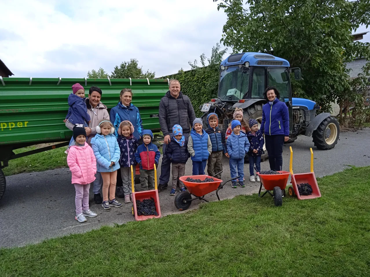 Eine Gruppe von Menschen, einschließlich Erwachsener und Kinder, steht vor einem Traktor und einem grünen Container, mit Schubkarren voller schwarzer Trauben auf dem Gras.
