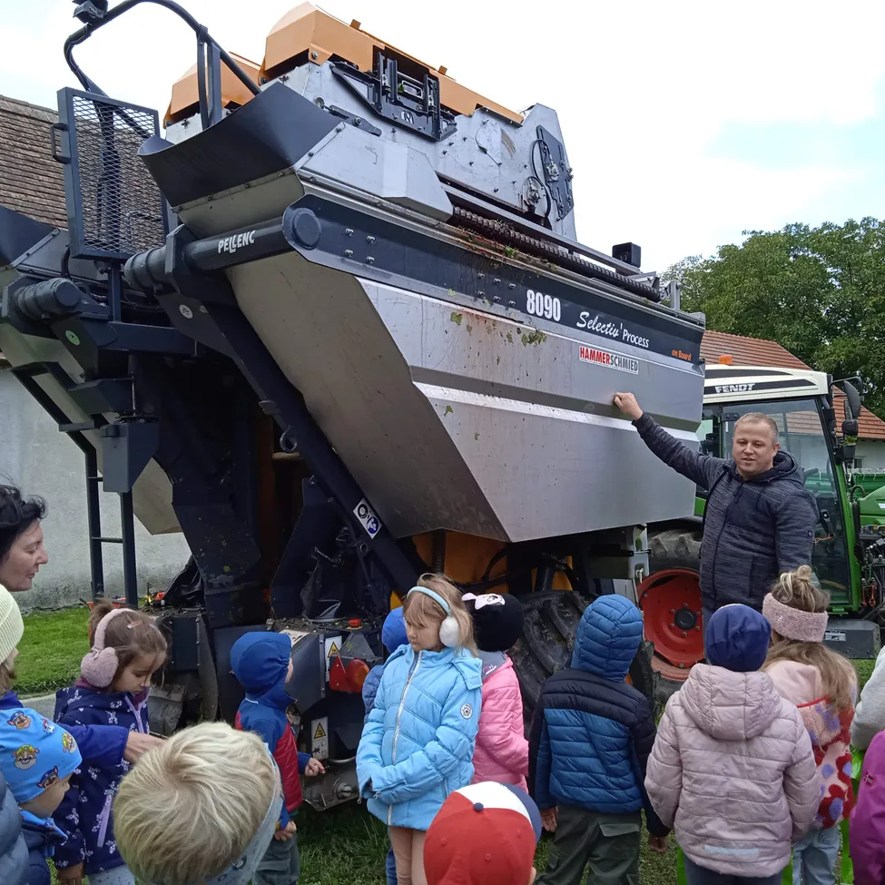 Ein Mann zeigt auf eine große landwirtschaftliche Maschine, während eine Gruppe von Kindern und Erwachsenen zusieht, alle warm gekleidet gegen das kalte Wetter.