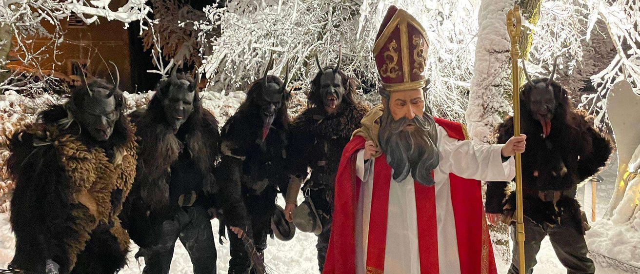 Eine Gruppe von Menschen in gruseligen Kostümen, einschließlich eines Bischofs, steht im Schnee. Sie halten Stöcke und scheinen eine Zeremonie durchzuführen.