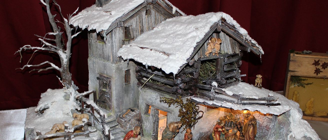 Ein detailliertes Modell einer Krippenszene mit einem verschneiten Dorf im Hintergrund. Die Figuren umfassen Maria, Josef und das Jesuskind, zusammen mit Hirten und Schafen. Die Szene ist unter einem Holzdach aufgebaut.
