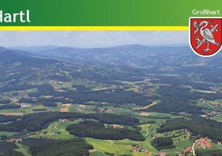Luftaufnahme von Gemeinde Hartl mit üppigen grünen Hügeln, einer entfernten Bergkette und drei Wappen oben.