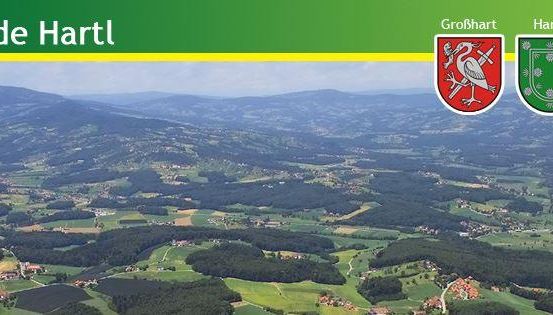 Luftaufnahme von Gemeinde Hartl mit üppigen grünen Hügeln, einer entfernten Bergkette und drei Wappen oben.