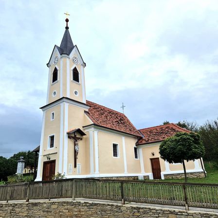 Eine kleine Kirche mit einem Turm, zwei Uhren und einem Kreuz darüber. Die Kirche hat ein rotes Dach und weiße Begrenzungen.