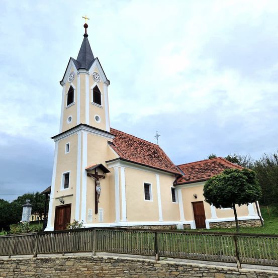 Eine kleine Kirche mit einem Turm, zwei Uhren und einem Kreuz darüber. Die Kirche hat ein rotes Dach und weiße Begrenzungen.
