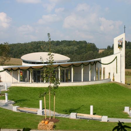 Ein modernes weißes Gebäude mit einem geschwungenen Dach und einem Glockenturm ist von einem üppigen grünen Rasen umgeben. Das Gebäude hat einen verglasten Eingang mit einer hölzernen Bank in der Nähe. Im Vordergrund sind kleine Bäume gepflanzt, mit Kürbissen auf dem Boden. In der Ferne gibt es Bäume und ein weites Feld.