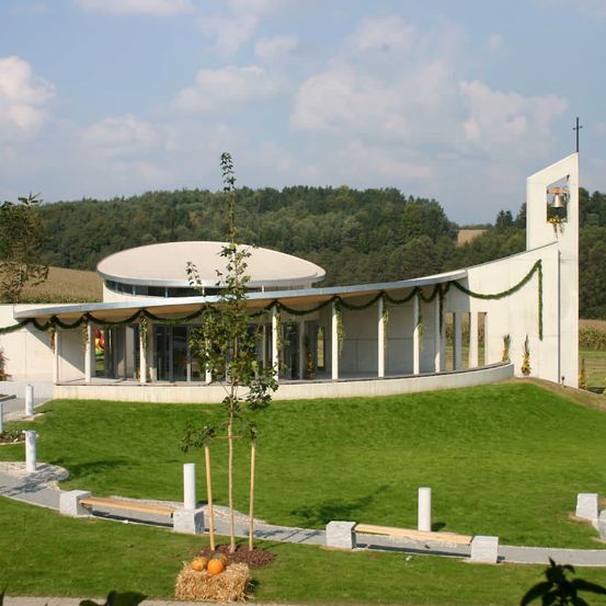 Ein modernes weißes Gebäude mit einem geschwungenen Dach und einem Glockenturm ist von einem üppigen grünen Rasen umgeben. Das Gebäude hat einen verglasten Eingang mit einer hölzernen Bank in der Nähe. Im Vordergrund sind kleine Bäume gepflanzt, mit Kürbissen auf dem Boden. In der Ferne gibt es Bäume und ein weites Feld.