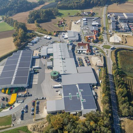 Luftaufnahme eines Industriekomplexes mit mehreren Gebäuden, Solarpanelen und einem von Bäumen und Feldern umgebenen Parkplatz.