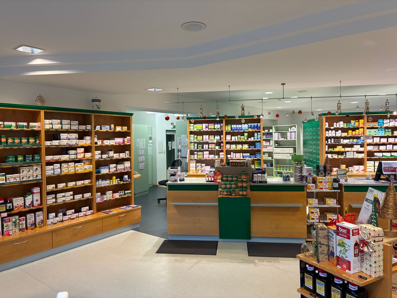 Eine Apotheke mit Regalen voller Produkte, einem Schreibtisch mit verschiedenen Artikeln und einem modernen Deckendesign. Der Boden ist weiß und sauber.