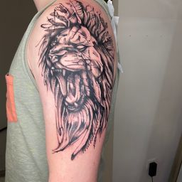 Ein Nahaufnahme eines Löwenkopf-Tattoos mit detaillierter Schattierung auf der Schulter einer Person, die ein graues Tanktop trägt.