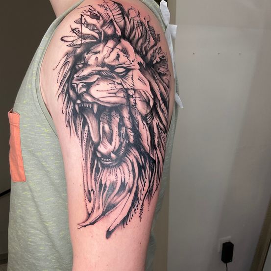 Ein Nahaufnahme eines Löwenkopf-Tattoos mit detaillierter Schattierung auf der Schulter einer Person, die ein graues Tanktop trägt.