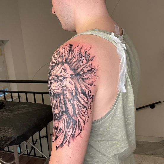 Ein Mann wird von hinten gezeigt und trägt ein großes Tattoo eines Löwenkopfes auf seiner linken Schulter. Er trägt ein ärmelloses grünes Shirt. Hinter ihm bedeckt ein schwarzer Stoff einen Stuhl, und ein schwarzes Geländer ist sichtbar.