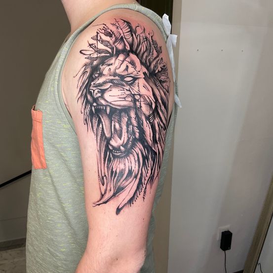 Ein Mann in einem ärmellosen grauen Shirt zeigt ein Löwentattoo am Oberarm. Das Tattoo ist schwarz-weiß und zeigt einen brüllenden Löwen.